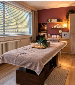 Table de massage drapée de serviettes avec plateau de bougies et plante verte dans une cabine de massage aux murs bordeaux, institut de beauté à Genève